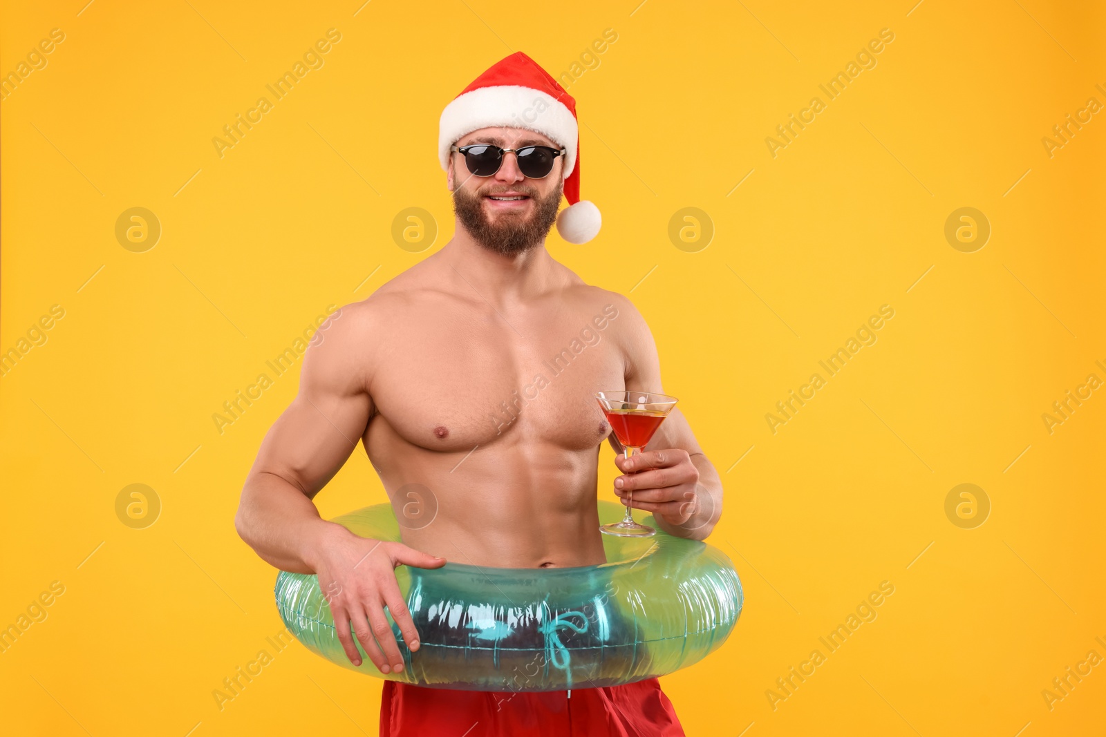 Photo of Muscular young man in Santa hat holding inflatable ring and cocktail on orange background. Space for text