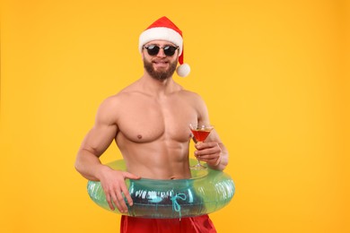 Muscular young man in Santa hat holding inflatable ring and cocktail on orange background. Space for text