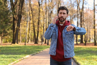 Emotional man talking by smartphone and checking time on watch in park, space for text. Being late concept