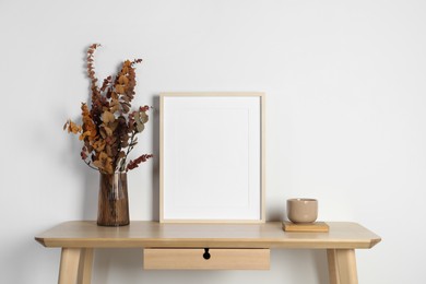 Photo of Empty photo frame, cup and vase with dry decorative leaves on wooden table. Mockup for design