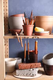 Photo of Set of different crafting tools and clay dishes on wooden rack in workshop