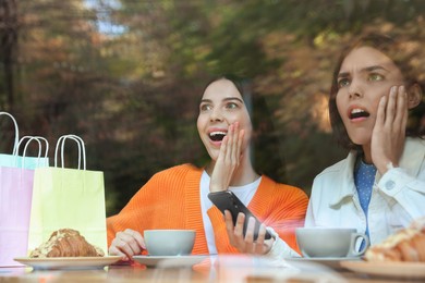 Special Promotion. Emotional young women using smartphone at table in cafe, view from outdoors