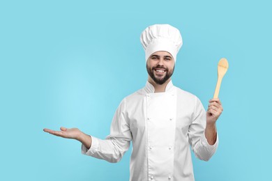 Photo of Happy young chef in uniform holding wooden spoon on light blue background