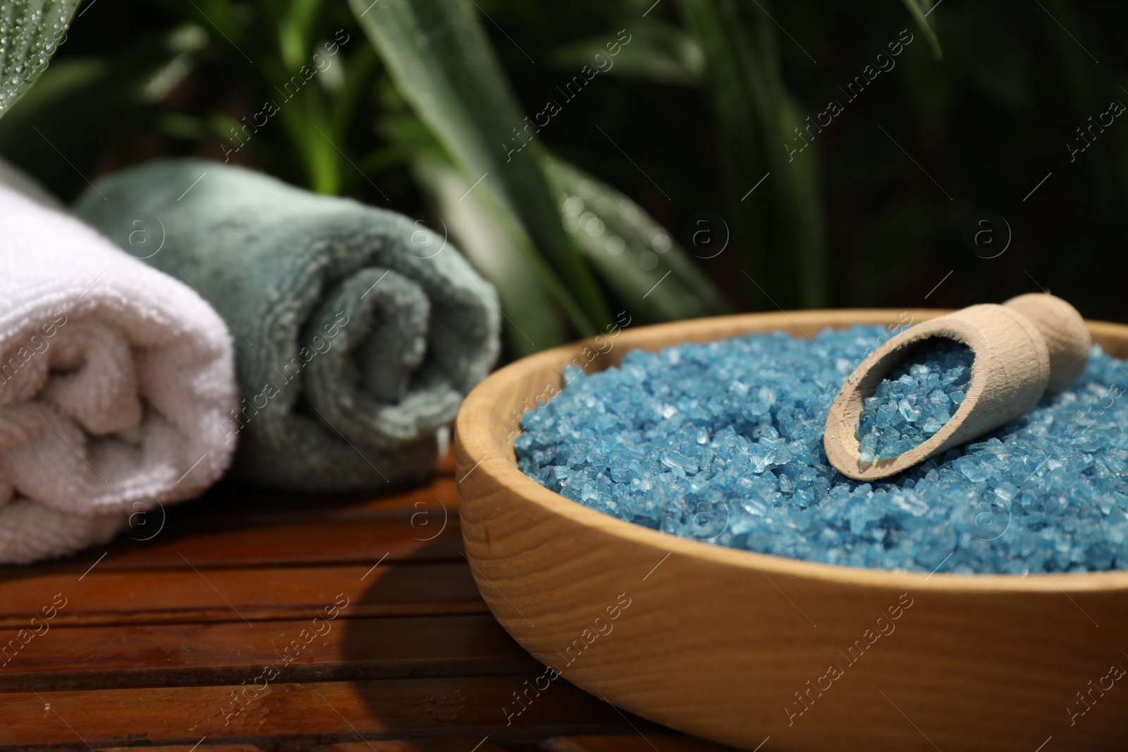 Photo of Bowl of blue sea salt with scoop and rolled towels on wooden table, closeup