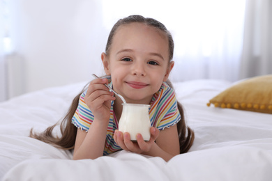 Photo of Cute little girl with tasty yogurt on bed at home