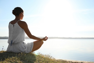 Young woman meditating near river at sunset, space for text. Nature healing power