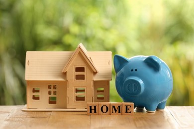 Photo of Piggy bank, house model and word Home made of cubes on wooden table outdoors