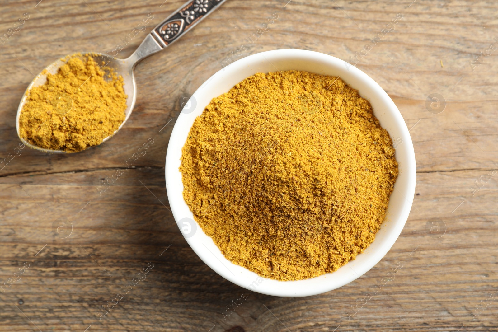 Photo of Dry curry powder in bowl and spoon on wooden table, top view