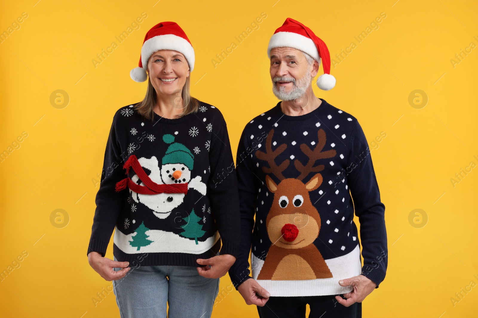 Photo of Happy senior couple in Santa hats showing Christmas sweaters on orange background