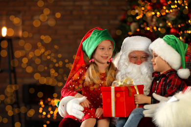 Photo of Santa Claus and little children with present near Christmas tree indoors