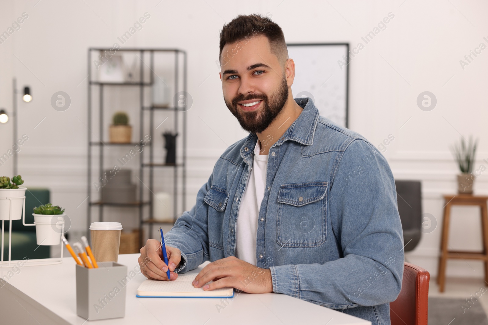 Photo of Young man writing in notebook at white table indoors