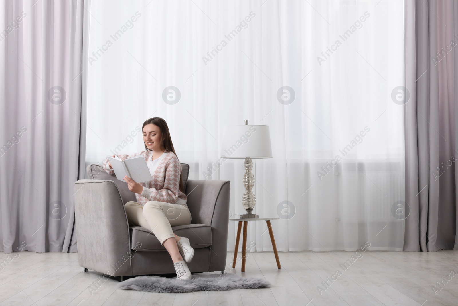 Photo of Woman reading book in armchair near window with stylish curtains at home. Space for text