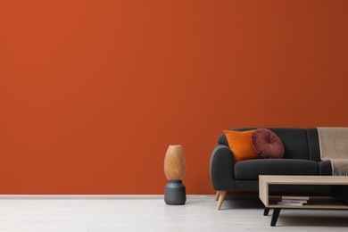 Photo of Stylish grey sofa with colorful pillows, wooden table and vase near dark orange wall indoors, space for text. Interior design