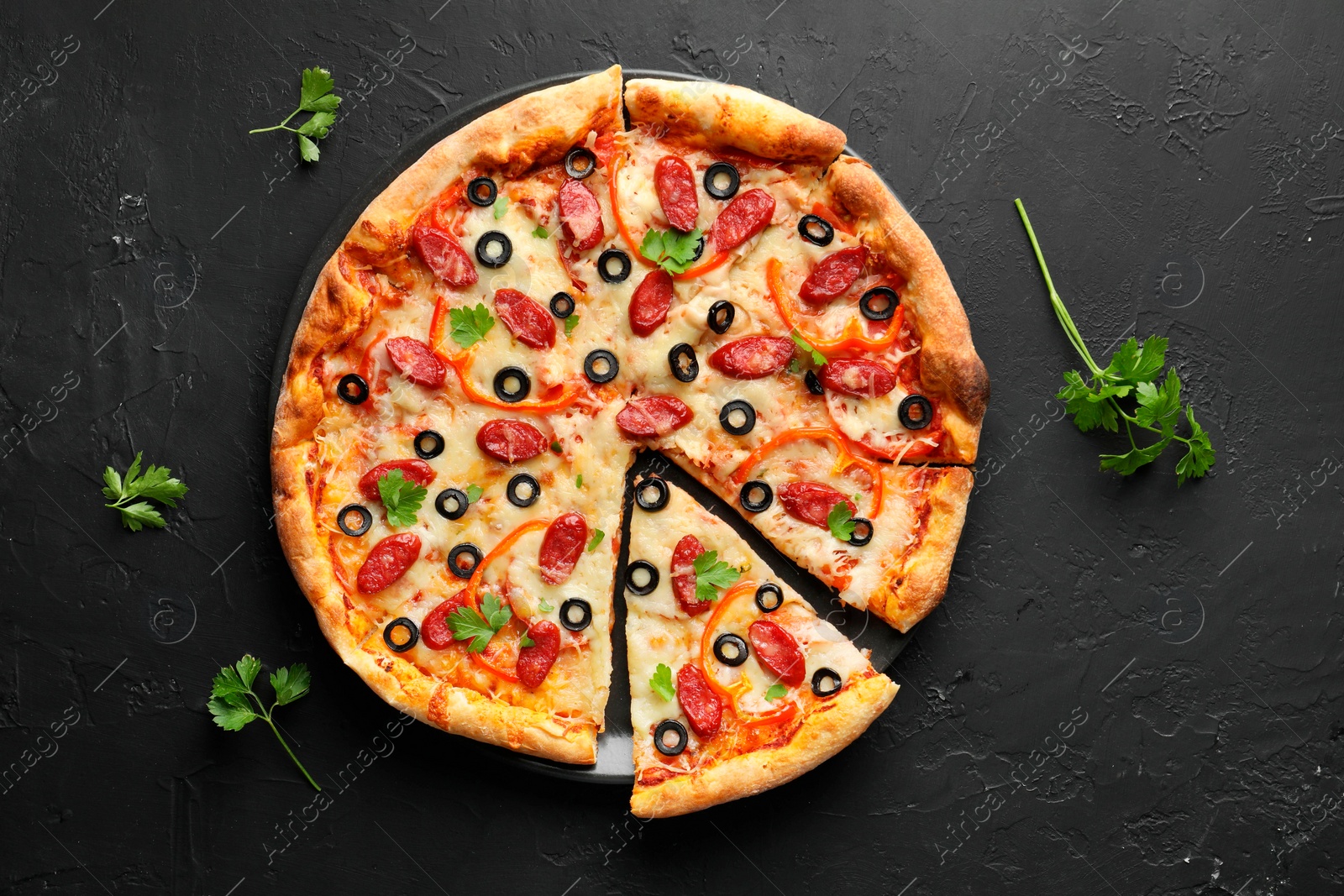 Photo of Tasty pizza with dry smoked sausages, olives, pepper and parsley on black table, top view