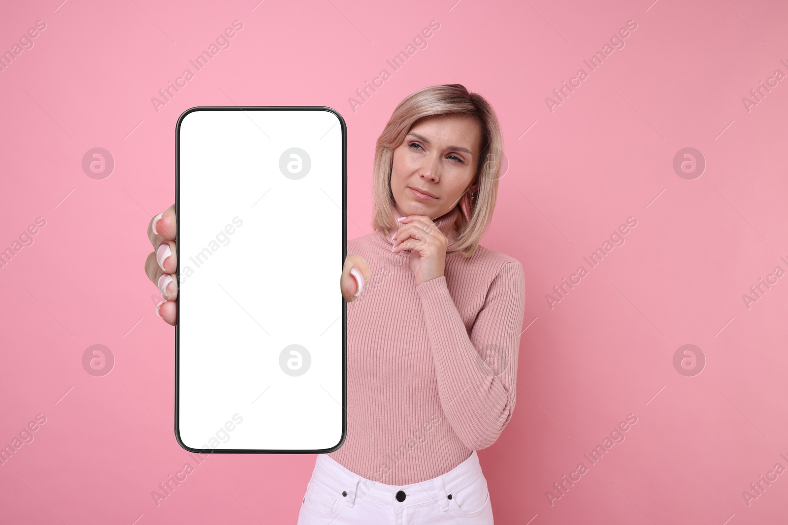 Image of Thoughtful woman showing mobile phone with blank screen on pink background. Mockup for design