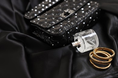 Photo of Leather bag, bottle of perfume and golden bracelets on black fabric, closeup. Space for text