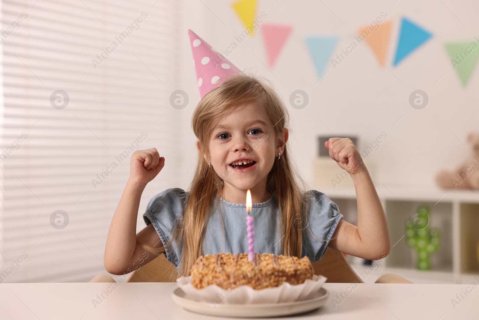 Photo of Cute girl in party hat with birthday cake at table indoors