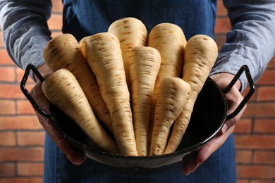 Photo of Woman holding bowl with delicious fresh ripe parsnips near red brick wall, closeup