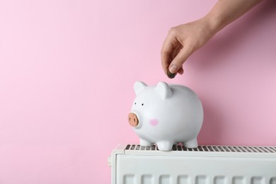 Photo of Woman putting coin into piggy bank on heating radiator against pink background, closeup. Space for text