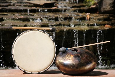 Photo of Drum and handpan with mallets near waterfall outdoors on sunny day. Percussion musical instruments