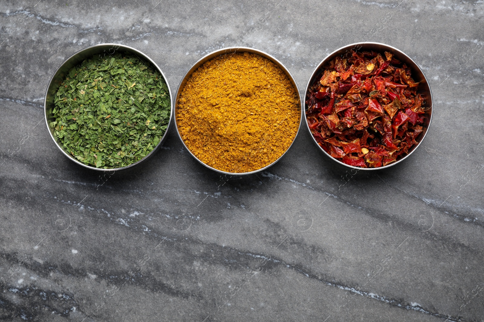 Photo of Bowls with dry parsley, turmeric and chili pepper on grey marble table, flat lay. Space for text