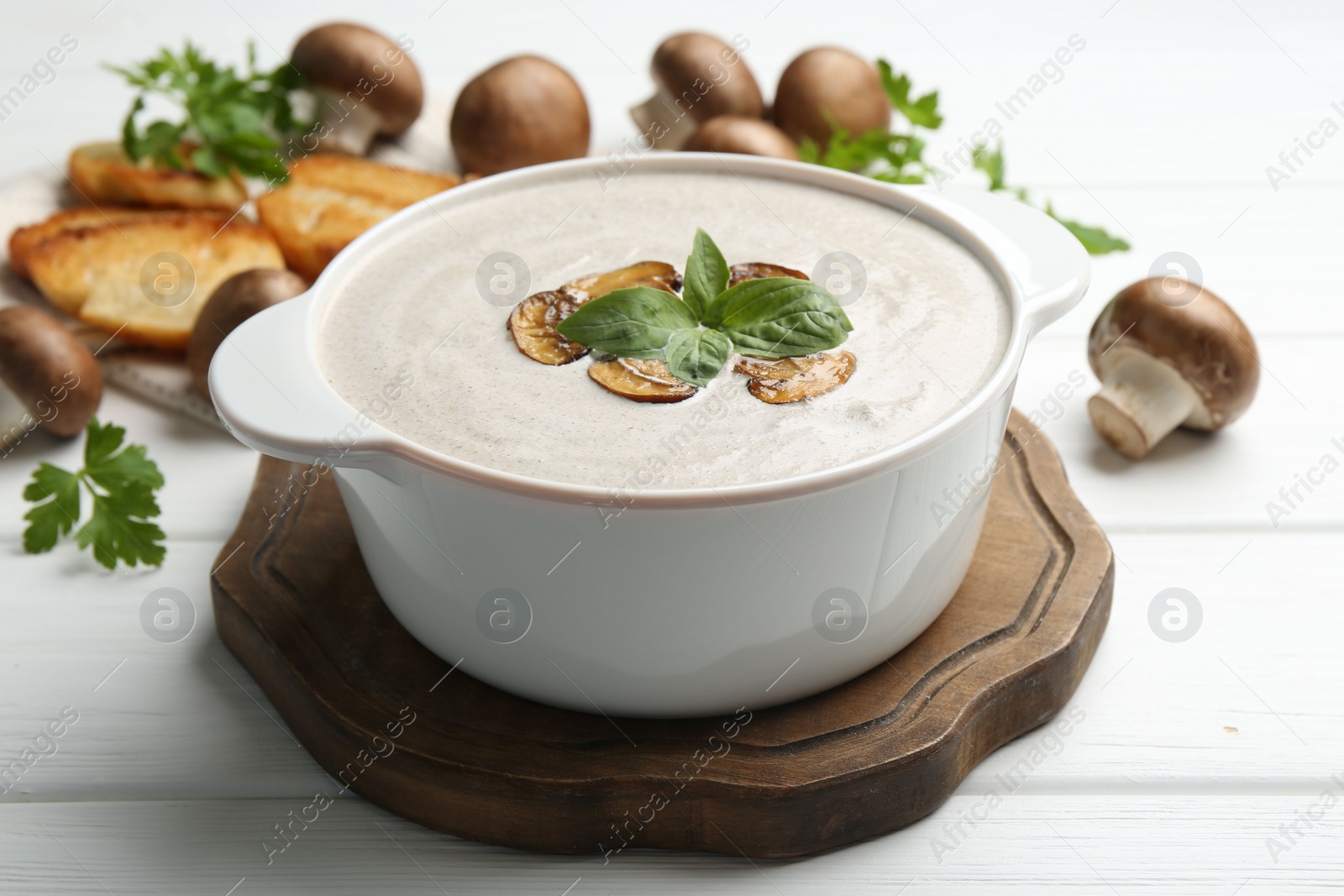 Photo of Delicious homemade mushroom soup in ceramic pot, croutons, fresh champignons and parsley on white wooden table