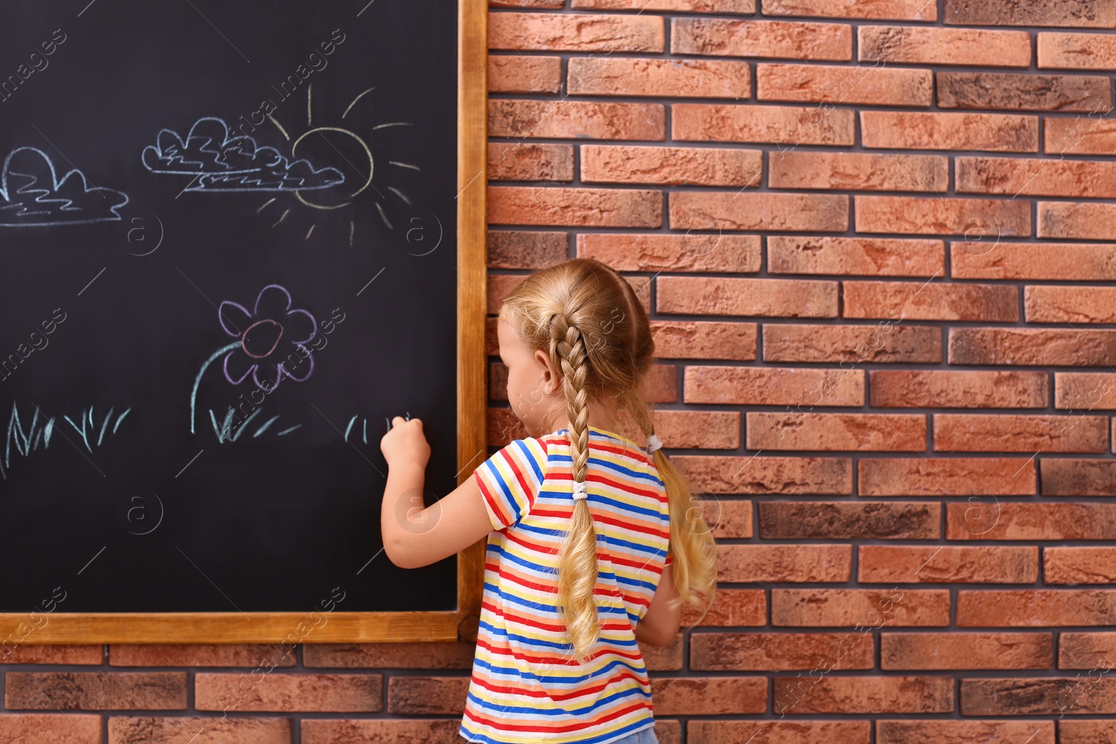 Photo of Cute little left-handed girl drawing on chalkboard near brick wall