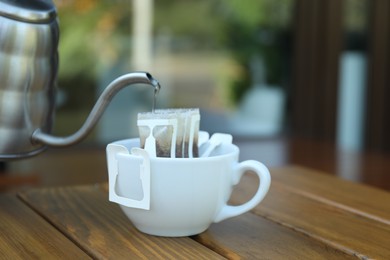 Pouring hot water into cup with drip coffee bag from kettle on wooden table, closeup