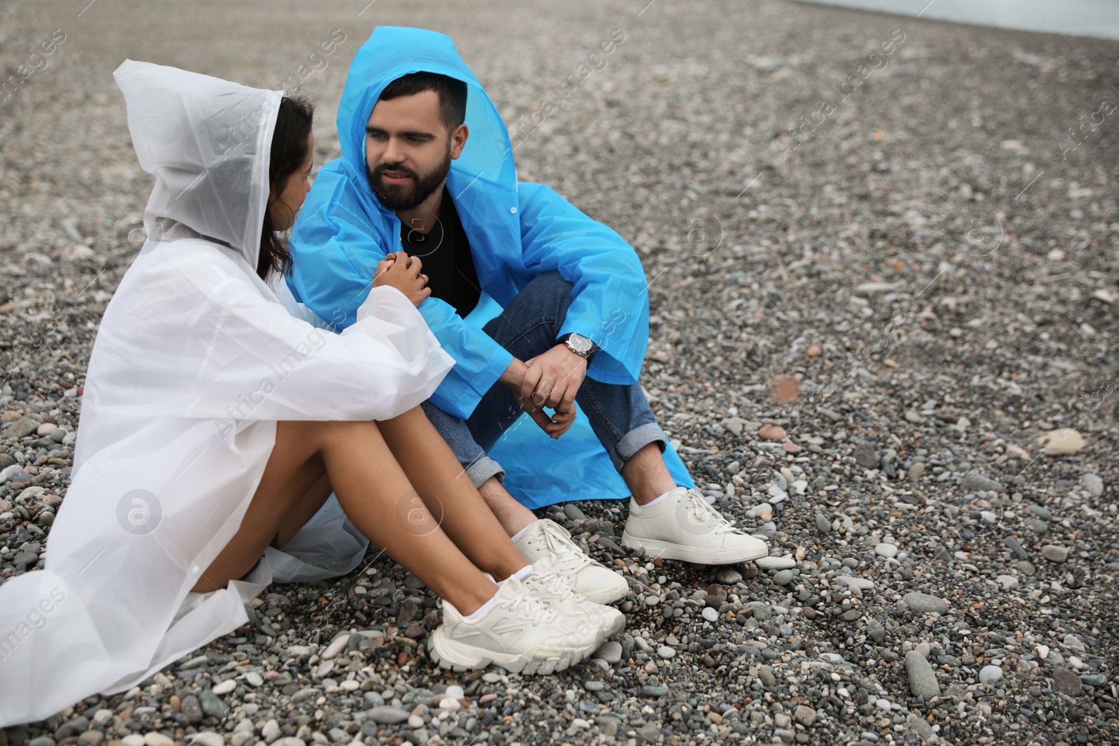 Photo of Young couple in raincoats enjoying time together under rain on beach, space for text