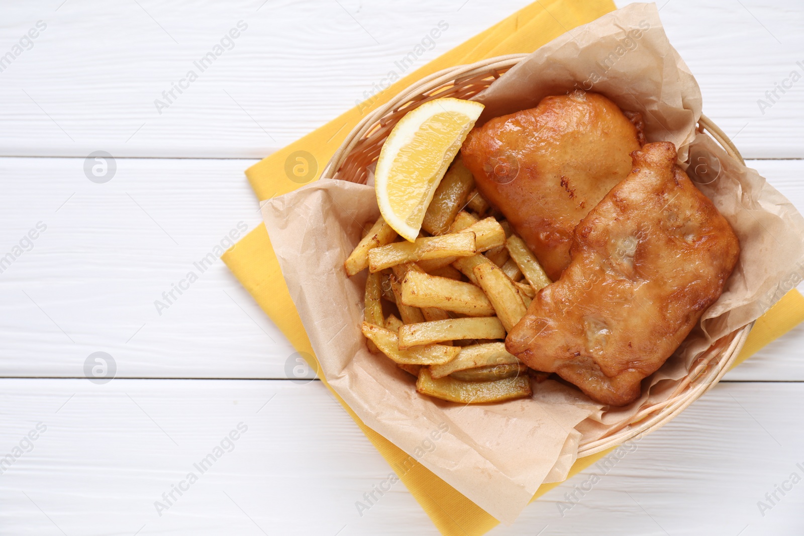 Photo of Tasty fish, chips and lemon in wicker bowl on white wooden table, top view. Space for text