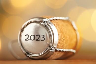 Image of Cork of sparkling wine and muselet cap with engraving 2023 on wooden table, closeup. Bokeh effect