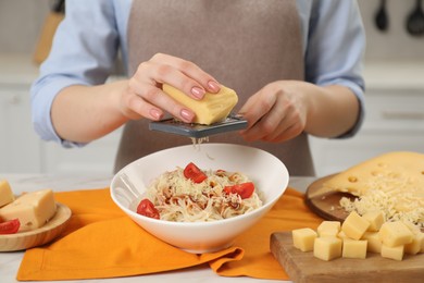 Photo of Woman grating cheese onto delicious pasta at white table in kitchen, closeup