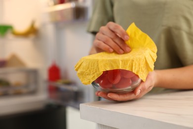Woman covering bowl of fresh tomatoes with beeswax food wrap at table in kitchen, closeup. Space for text