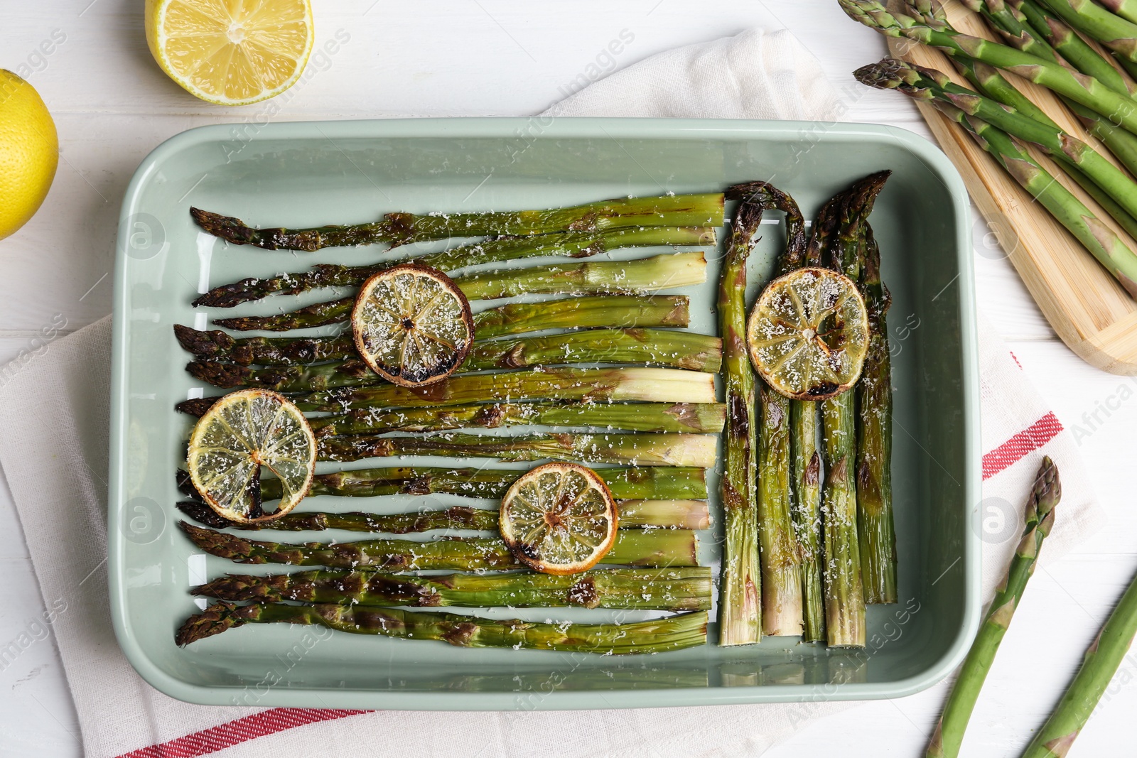 Photo of Oven baked asparagus with lemon slices in ceramic dish on white table, flat lay