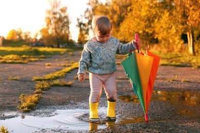 Photo of Little girl wearing rubber boots with colorful umbrella in puddle outdoors, space for text. Autumn walk