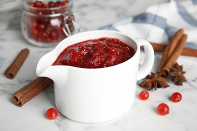 Photo of Pitcher of cranberry sauce and spices on marble table, closeup
