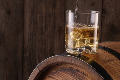 Pouring whiskey into glass on barrel against wooden background, closeup. Space for text