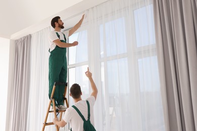 Photo of Workers in uniform hanging window curtain indoors, low angle view