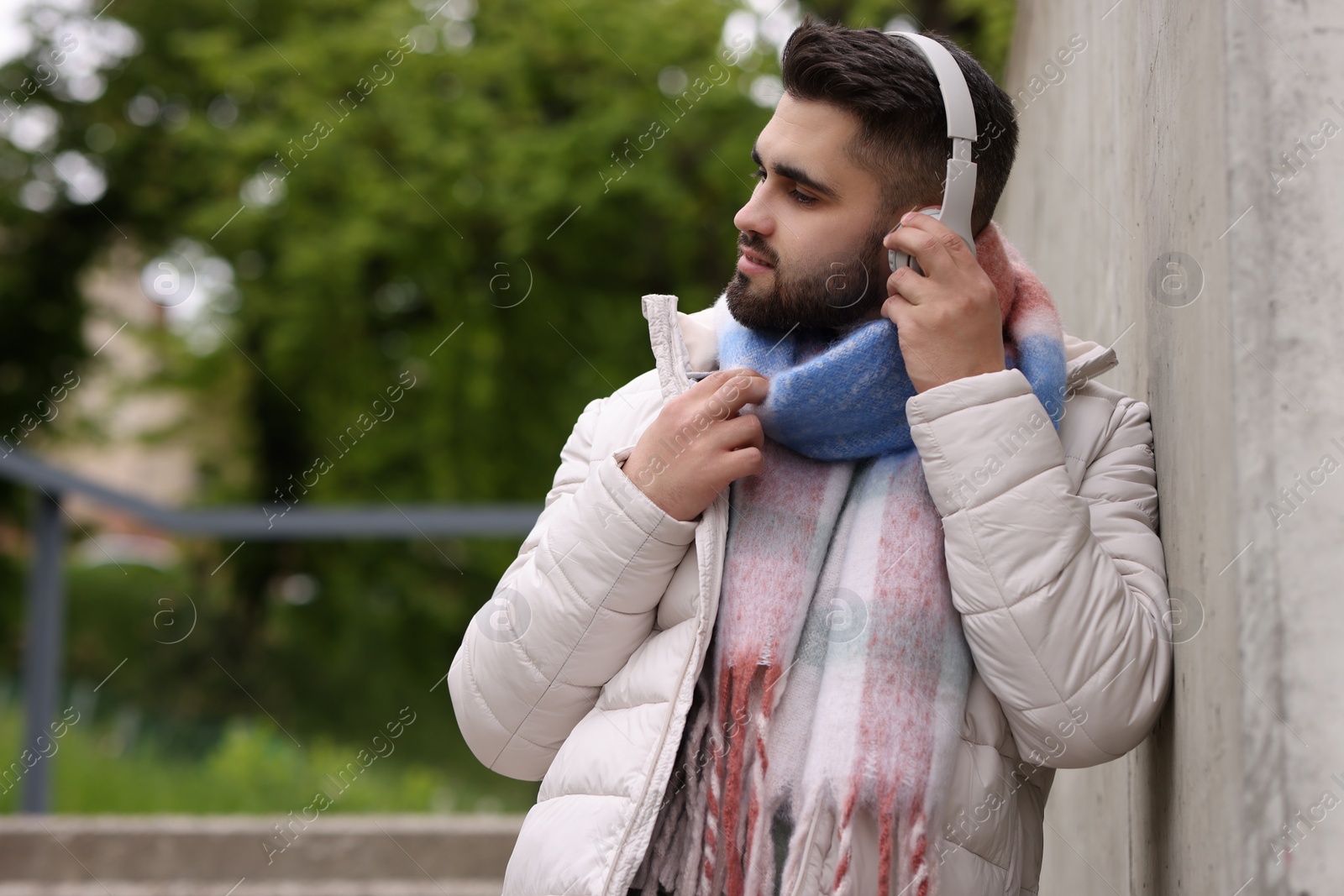 Photo of Handsome man in warm scarf and headphones near wall on city street. Space for text