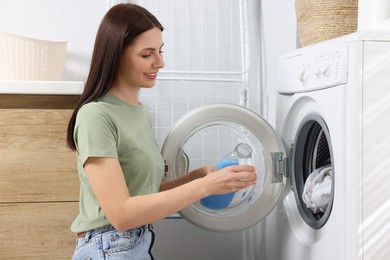 Photo of Woman pouring fabric softener from bottle into cap near washing machine in bathroom