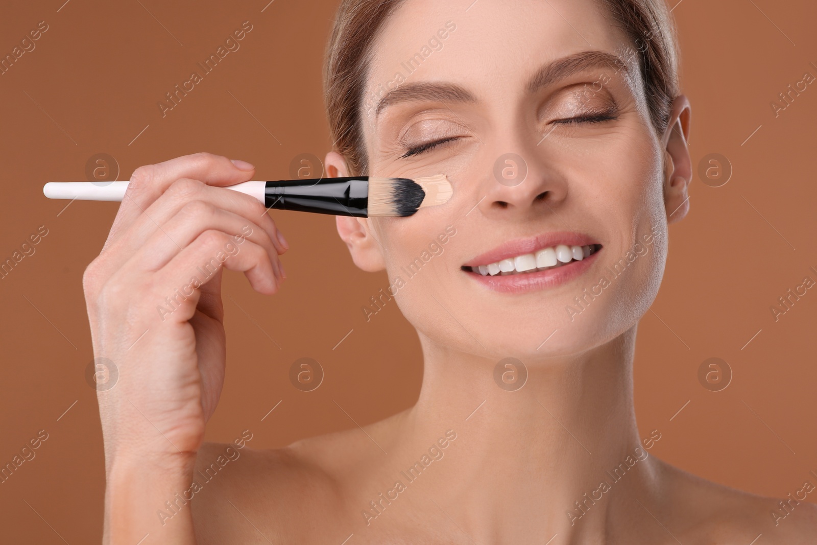 Photo of Woman applying foundation on face with brush against brown background