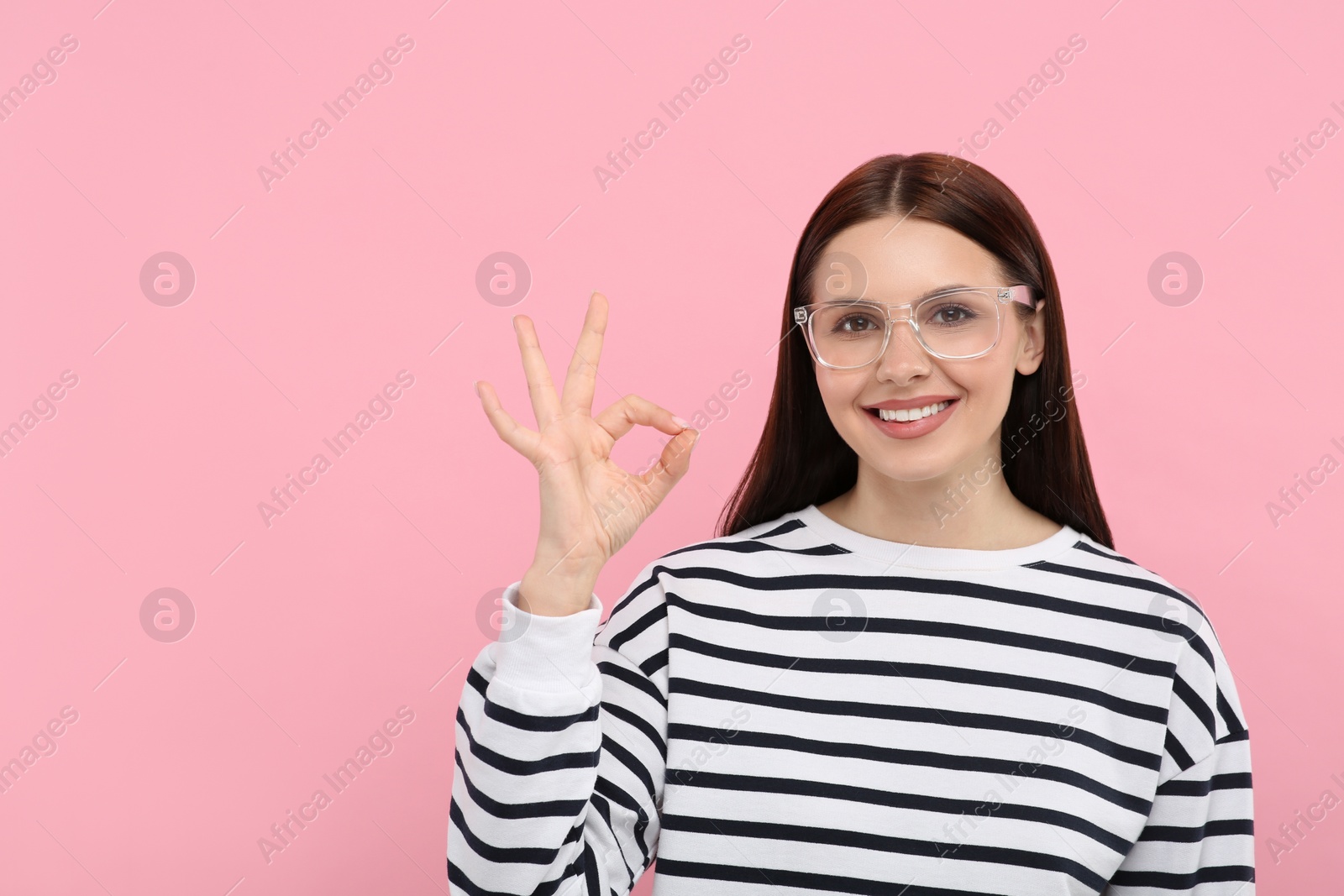Photo of Smiling woman in stylish eyeglasses showing ok gesture on pink background. Space for text