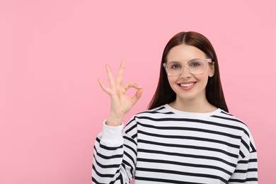 Smiling woman in stylish eyeglasses showing ok gesture on pink background. Space for text