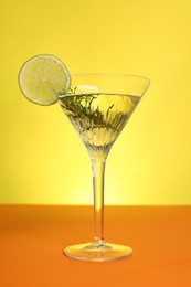 Photo of Martini glass of refreshing cocktail with lemon slice and rosemary on orange table