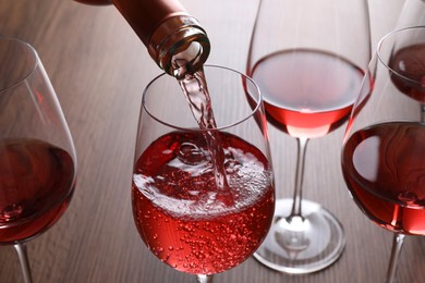 Photo of Pouring rose wine from bottle into glasses on wooden table, closeup