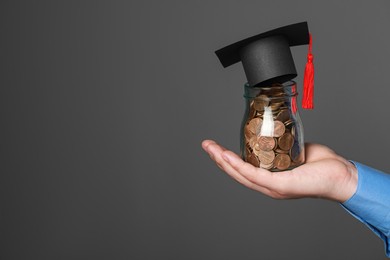Photo of Man holding glass jar of coins and graduation cap against dark grey background, closeup with space for text. Scholarship concept