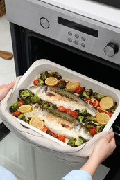 Photo of Woman taking baking dish with delicious fish and vegetables from oven, closeup