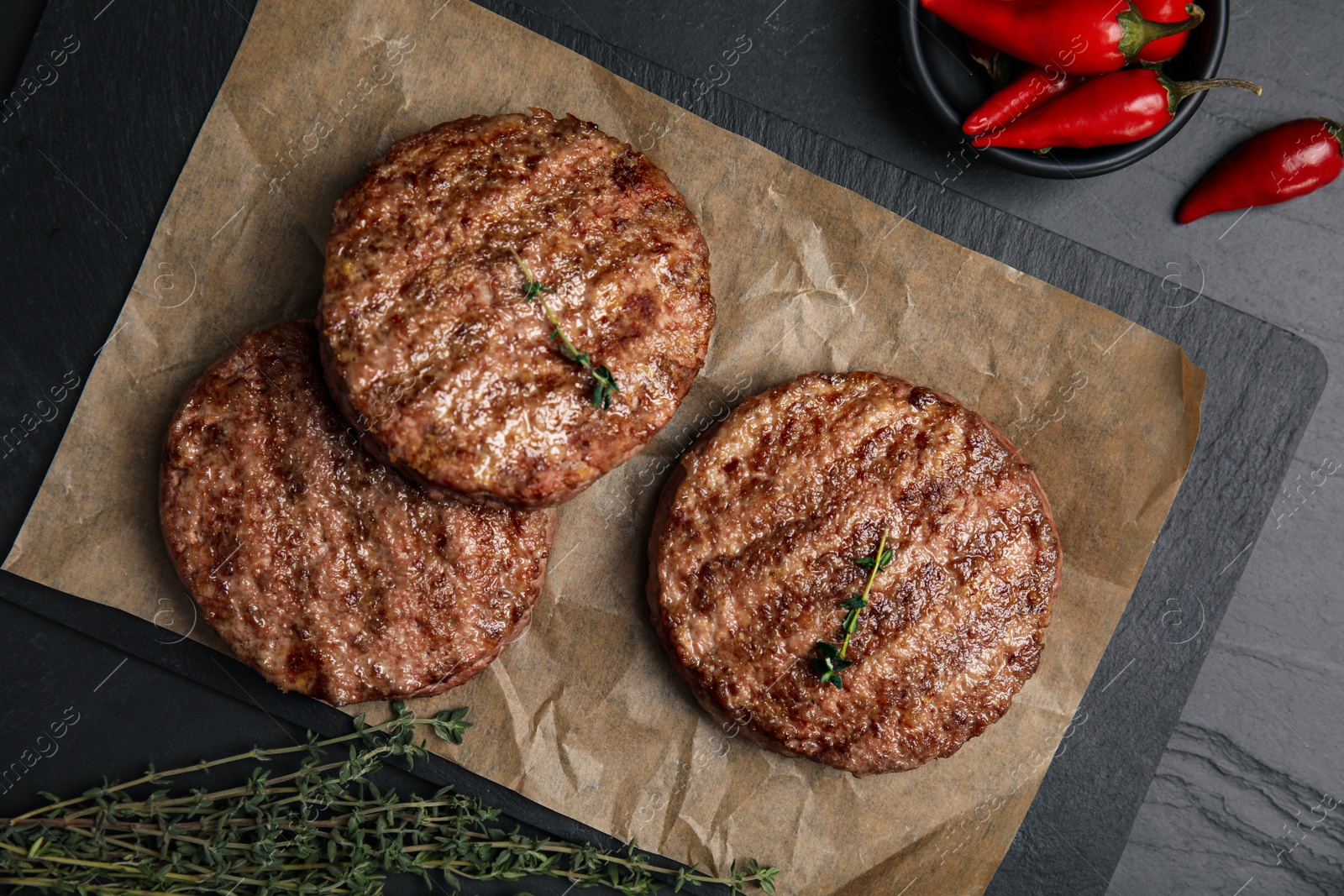 Photo of Tasty grilled hamburger patties, thyme and chili peppers on black table, flat lay