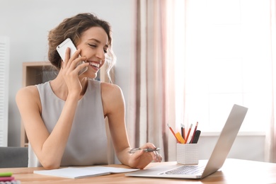 Photo of Young woman talking on mobile phone while working with laptop at desk. Home office