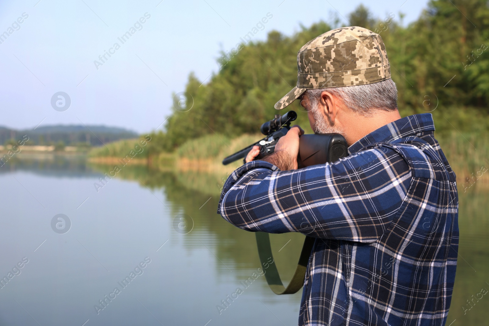 Photo of Man aiming with hunting rifle near lake outdoors. Space for text